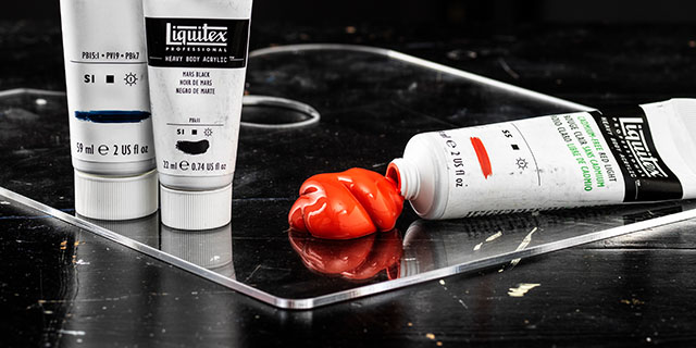 En bild med tre tuber Liquitex Heavy Body akrylfärg - varav en ligger ner med röd färg som kommer ut ur tuben. 