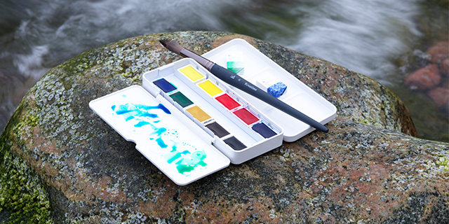 Bild på ett akvarellset från Winsor & Newton som ligger på en sten vid vatten